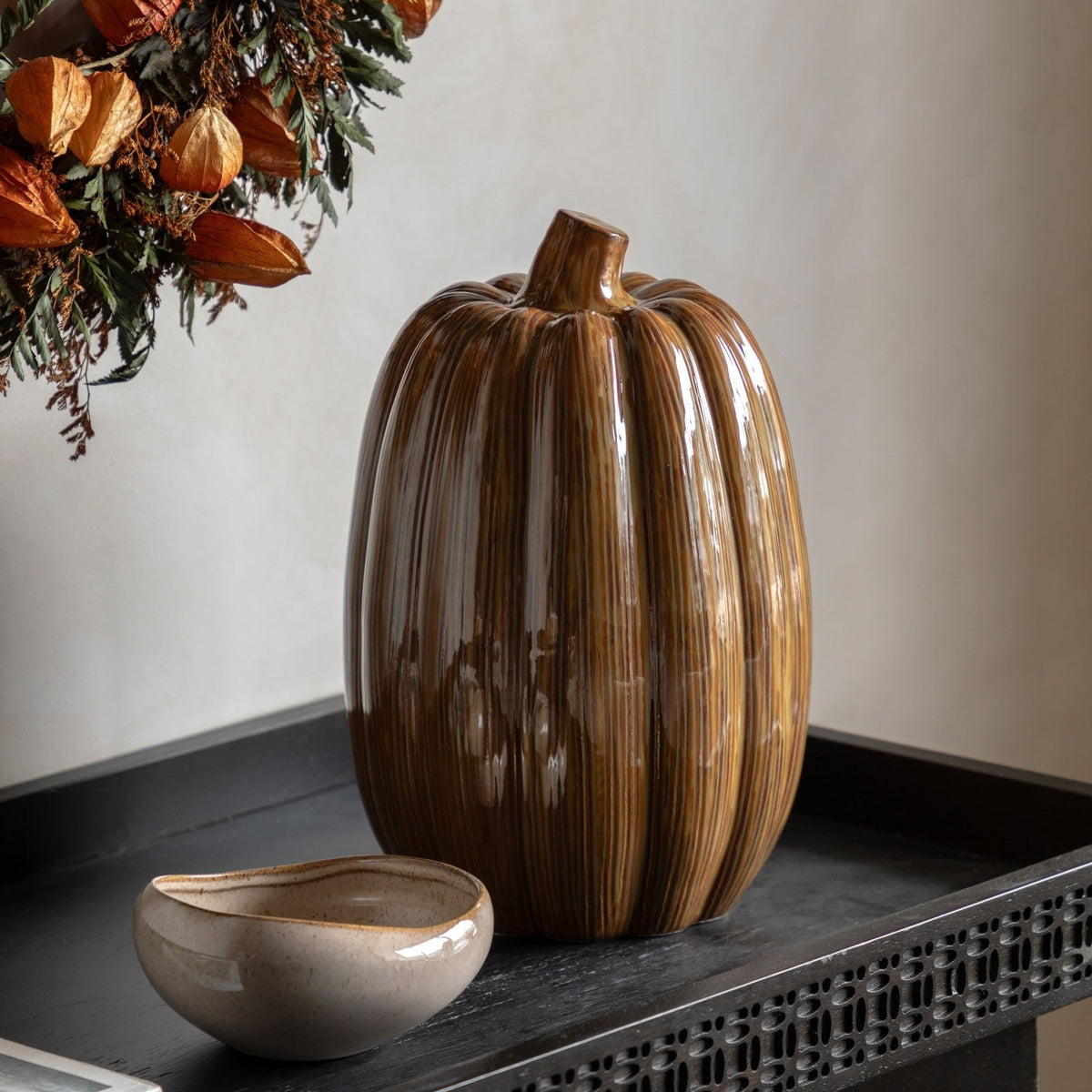 Decorative ceramic pumpkin ornament with glossy striped glaze styled on a black console table with autumn wreath and stoneware bowl – modern seasonal home décor.