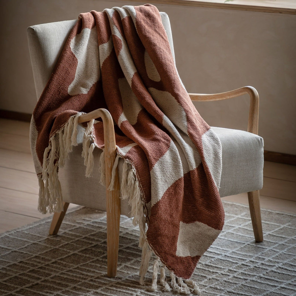 Rust and cream patterned throw blanket with tassel fringe draped over an armchair in a cosy living room setting.