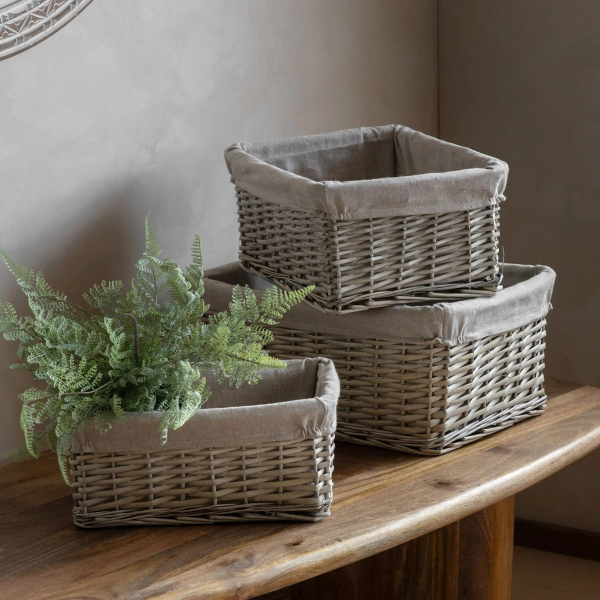 Set of three grey wicker storage baskets with linen lining, displayed on wooden table with decorative plant, perfect for home organisation and rustic interiors.