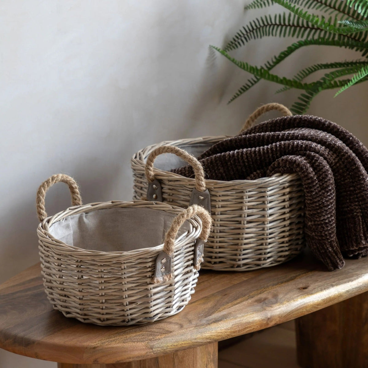 Set of two round wicker storage baskets with rope handles, styled on a wooden bench with a cosy brown throw blanket.