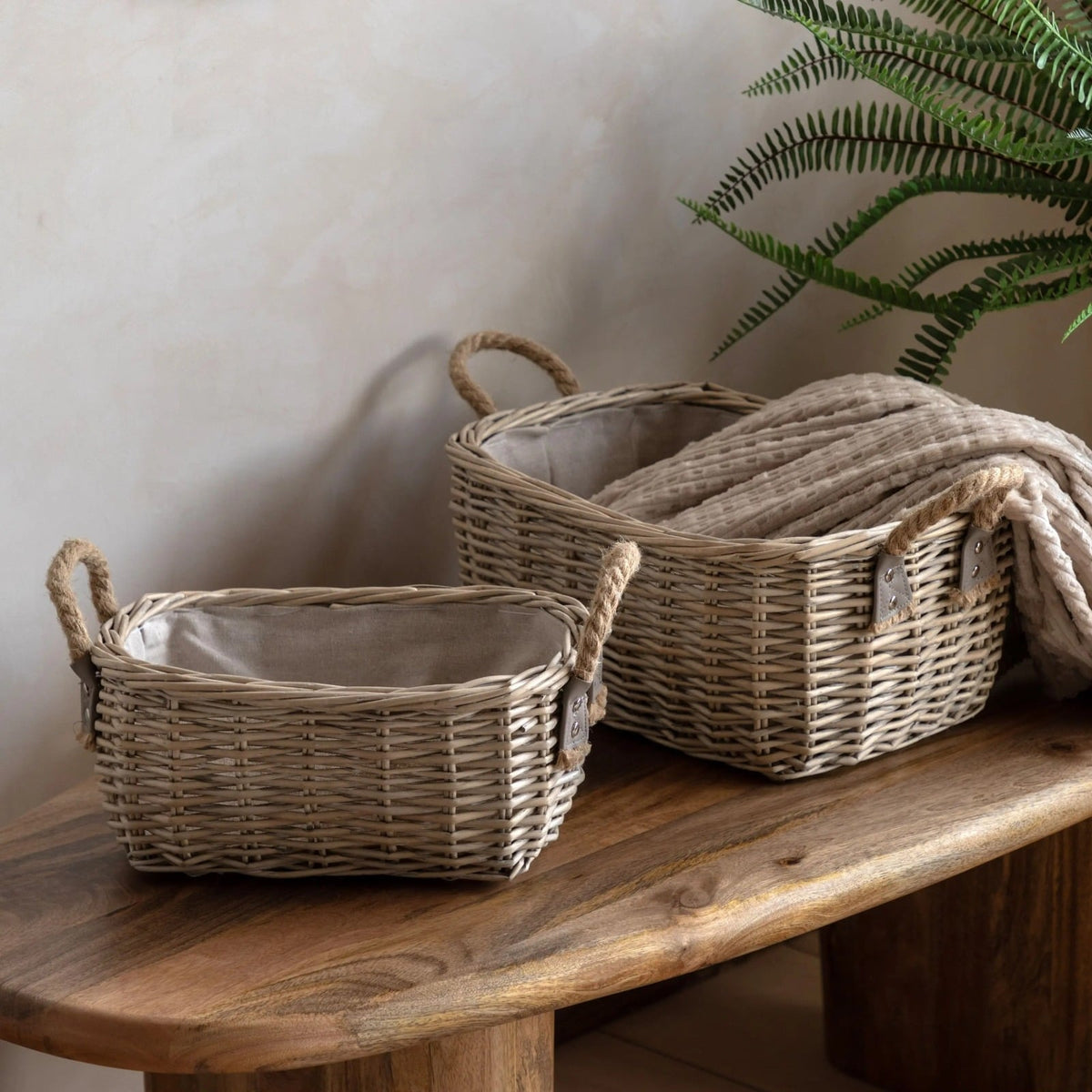 Two rectangular wicker storage baskets with rope handles and linen lining, styled on a wooden bench beside a green plant.