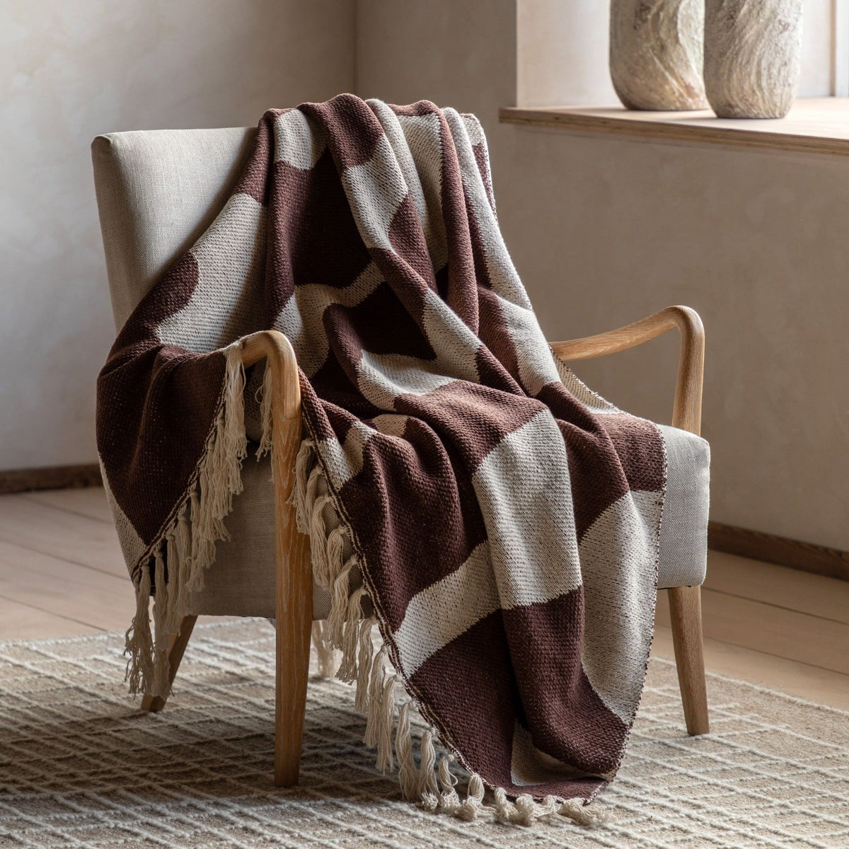 Brown and cream patterned throw blanket with tassel edging draped over a modern wooden armchair in a minimalist living room.