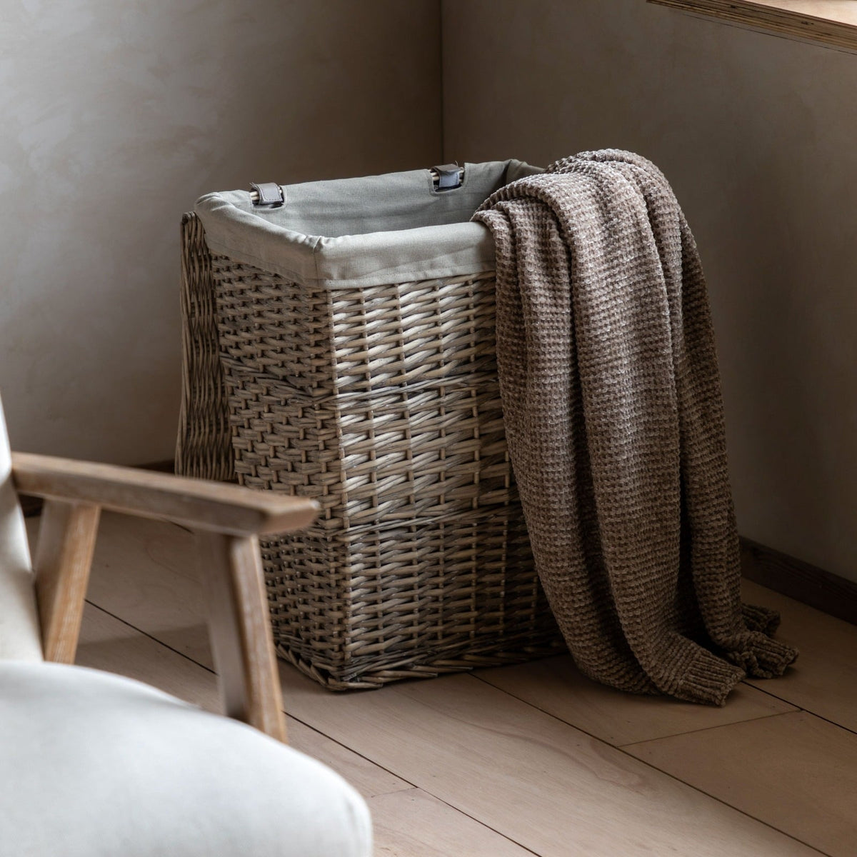 Natural wicker laundry basket with fabric lining and textured throw, styled in a cosy bedroom corner.