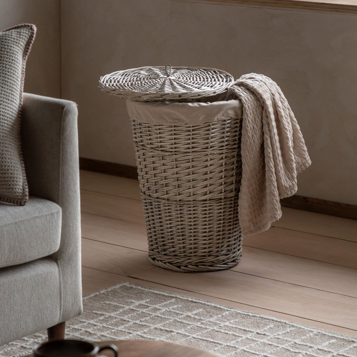 Grey wicker laundry basket with fabric liner and lid, styled in a neutral living room with a beige throw draped over the side.